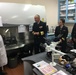 Three U.S. Navy personnel in dress uniforms are visiting a laboratory, appearing to receive a briefing or tour from a civilian staff member wearing a white lab coat. The setting is a scientific laboratory identified as a Parasitology Department, equipped with microscopes, a biosafety cabinet, and supply storage. The interaction suggests an official inspection or command visit to a military medical or research facility.