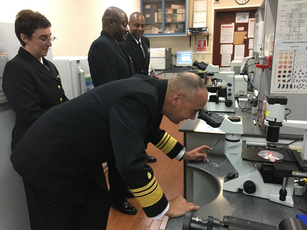 Four individuals in dark naval/military dress uniforms are gathered in a laboratory setting. One senior officer, identifiable by gold sleeve stripes indicating high rank, is bent over and looking through an Olympus microscope. Three other uniformed personnel observe from behind, suggesting an official inspection or demonstration visit.