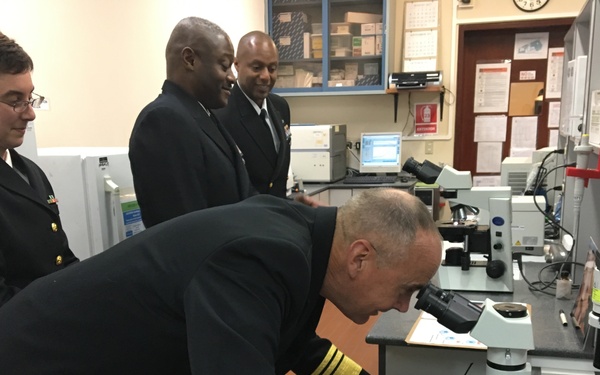 Four individuals in dark naval/military dress uniforms are gathered in a laboratory setting. One senior officer, identifiable by gold sleeve stripes indicating high rank, is bent over and looking through an Olympus microscope. Three other uniformed personnel observe from behind, suggesting an official inspection or demonstration visit.