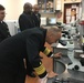 Four individuals in dark naval/military dress uniforms are gathered in a laboratory setting. One senior officer, identifiable by gold sleeve stripes indicating high rank, is bent over and looking through an Olympus microscope. Three other uniformed personnel observe from behind, suggesting an official inspection or demonstration visit.