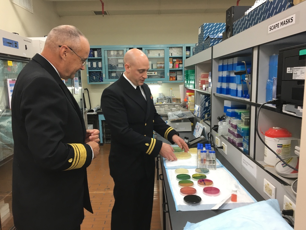 Two naval officers in dark dress uniforms with gold sleeve stripes are examining laboratory materials in what appears to be a military or government research laboratory. One officer is pointing at a collection of colorful petri dishes and test tube racks on a lab bench. The setting includes shelving with laboratory supplies and a label reading 'SCAPE MASKS' visible in the upper right.