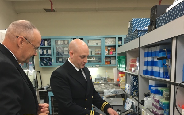 Two naval officers in dark dress uniforms with gold sleeve stripes are examining laboratory materials in what appears to be a military or government research laboratory. One officer is pointing at a collection of colorful petri dishes and test tube racks on a lab bench. The setting includes shelving with laboratory supplies and a label reading 'SCAPE MASKS' visible in the upper right.