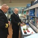 Two naval officers in dark dress uniforms with gold sleeve stripes are examining laboratory materials in what appears to be a military or government research laboratory. One officer is pointing at a collection of colorful petri dishes and test tube racks on a lab bench. The setting includes shelving with laboratory supplies and a label reading 'SCAPE MASKS' visible in the upper right.