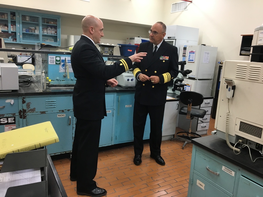 Two men in dark naval uniforms are engaged in conversation inside what appears to be a military medical or scientific laboratory. The officer on the right displays higher rank insignia including gold sleeve stripes and a row of service ribbons, while the officer on the left appears to be briefing or explaining something. The lab setting features teal/blue cabinetry, scientific equipment, and storage shelving typical of a military medical facility.
