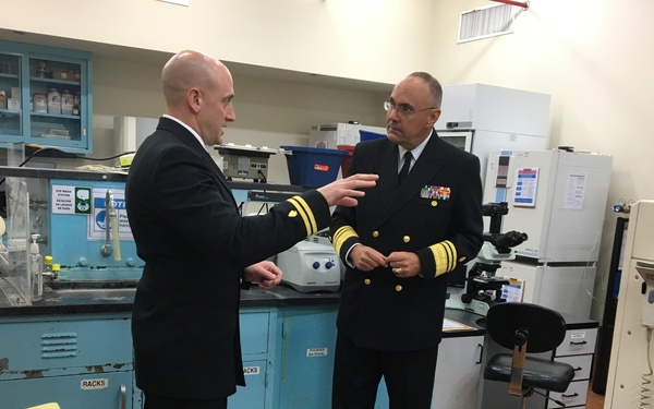 Two men in dark naval uniforms are engaged in conversation inside what appears to be a military medical or scientific laboratory. The officer on the right displays higher rank insignia including gold sleeve stripes and a row of service ribbons, while the officer on the left appears to be briefing or explaining something. The lab setting features teal/blue cabinetry, scientific equipment, and storage shelving typical of a military medical facility.