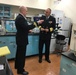 Two men in dark naval uniforms are engaged in conversation inside what appears to be a military medical or scientific laboratory. The officer on the right displays higher rank insignia including gold sleeve stripes and a row of service ribbons, while the officer on the left appears to be briefing or explaining something. The lab setting features teal/blue cabinetry, scientific equipment, and storage shelving typical of a military medical facility.