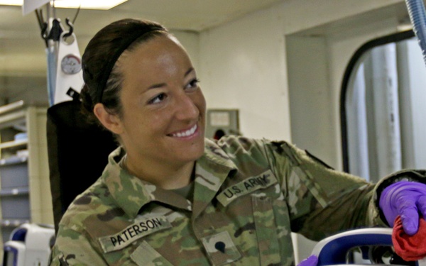 A U.S. Army soldier named Paterson is smiling while cleaning or maintaining a Zoll Propaq medical monitor/defibrillator device. She is wearing OCP (Operational Camouflage Pattern) uniform with purple nitrile gloves and using a red cloth to wipe down the equipment. The setting appears to be a military medical facility or ambulance interior with various medical equipment visible in the background.