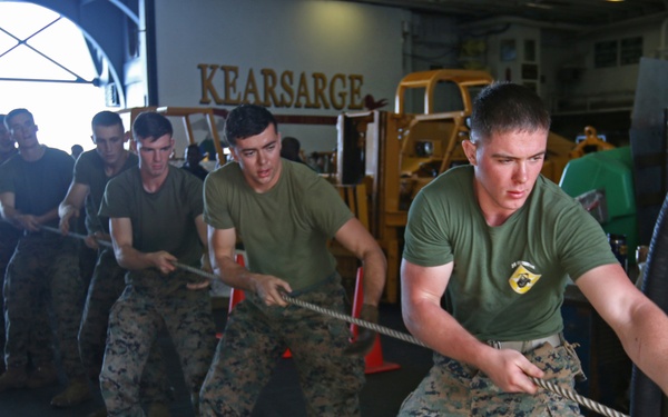 A group of U.S. Marines in military utility uniforms are engaged in a tug-of-war competition aboard the USS Kearsarge, an amphibious assault ship. The participants are pulling a thick rope in a hangar bay area, with the ship's name 'KEARSARGE' prominently displayed on the wall in the background. The activity appears to be a morale or physical fitness event conducted in the ship's vehicle/aircraft hangar deck.
