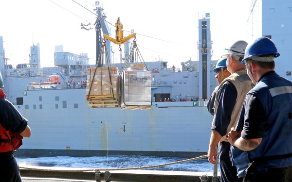 A replenishment-at-sea (RAS) operation is underway between two naval or military vessels. Crew members on the receiving ship's deck observe as a pallet of cargo is transferred via a highline/jackstay system suspended between the two ships. Personnel are wearing safety gear including hard hats and life vests.