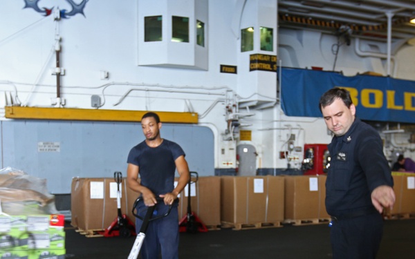 The image shows what appears to be the hangar bay of a naval vessel where personnel are conducting a stores or supply loading operation. Two sailors in dark navy uniforms are prominently visible, with one operating a red manual pallet jack to move a loaded pallet. Multiple pallets of boxed supplies are staged throughout the space.