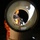 Preparing for a Disaster on Kauai A nighttime photograph taken looking into the open end of a large cylindrical pipe or tube. Through the pipe opening, a worker wearing high-visibility orange clothing is visible, along with what appears to be equipment or machinery mounted inside. Orange protective wrapping or sheeting is visible on the exterior of the pipe.
