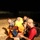 Preparing for a Disaster on Kauai A nighttime outdoor scene showing several individuals seated on dry grass, with one man in a red shirt and tan cap working on a laptop computer. A person in a bright orange/yellow hazmat or protective suit is visible in the background. The setting appears to be a field operation or training exercise.