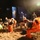Preparing for a Disaster on Kauai A nighttime emergency response scene showing multiple personnel, some wearing orange hazmat/chemical protective suits, gathered near command vehicles. The team appears to be in a rest or debriefing phase between operations, with some using laptops and communication devices. The scene is illuminated by artificial lighting from nearby vehicles and equipment.
