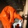 Preparing for a Disaster on Kauai Two individuals wearing bright orange hazmat/protective suits are moving through a narrow corridor or passageway inside what appears to be a metal-roofed industrial or temporary structure. The foreground figure is bending forward and reaching toward equipment mounted on a white structural post. A second similarly suited individual is visible further down the corridor.