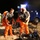 Preparing for a Disaster on Kauai Two personnel are donning or adjusting bright orange hazmat/chemical protective suits at night in an outdoor staging area. They are wearing tactical vests over the suits and appear to be preparing for or returning from a hazardous materials operation. A blue utility/response vehicle is visible in the background, along with a third person in a high-visibility yellow-green vest.