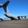 Utah's 85th CST transports equipment on a C-17 to Kauai A large military cargo aircraft, identified as a C-17 Globemaster III belonging to the Mississippi Air National Guard (ANG), is parked on a tarmac with its rear cargo ramp lowered. A blue ambulance or emergency vehicle is positioned near the open cargo bay, and two personnel in military uniforms are visible on the tarmac. The setting appears to be a coastal airfield under clear blue skies.