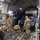 Utah's 85th CST load equipment on a C-17 to return from Kauai Military personnel in camouflage uniforms are securing vehicles inside the cargo hold of a large military transport aircraft, likely a C-17 Globemaster III. Multiple vehicles including what appears to be a blue ambulance/emergency vehicle and pickup trucks are being chained down to the aircraft floor. The scene depicts a standard military airlift loading operation with tie-down chains being attached to vehicle anchor points.