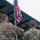 Two military personnel are photographed from behind, saluting an American flag on a flagpole. The soldier on the left wears a U.S. Army MultiCam (OCP) uniform with the name tape 'JORDAN' visible on his cap, while the soldier on the right wears a different pixelated camouflage uniform consistent with a non-U.S. military force. The setting appears to be an outdoor covered area or facility.