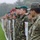 A multinational military formation is standing in a line outdoors on a grass field, likely during a joint exercise or ceremony. Soldiers from multiple nations are visible, with at least one Turkish soldier identifiable by the 'Türkiye' patch and Turkish flag on his uniform. Various military uniforms, camouflage patterns, and headgear styles indicate participation from several different countries.
