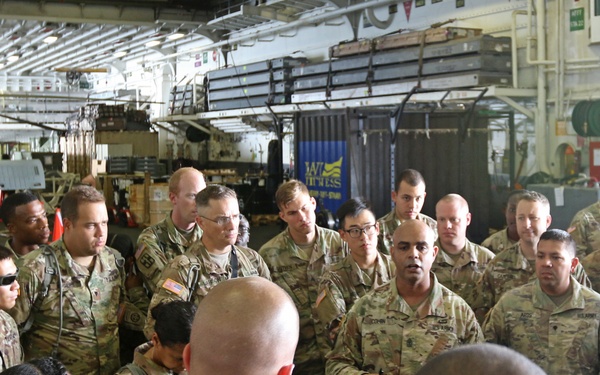 A large group of U.S. Army soldiers in MultiCam/OCP camouflage uniforms are gathered in what appears to be the vehicle deck or hangar bay of a military ship or large transport vessel. A briefing or formation appears to be taking place, with one soldier appearing to address the group. The setting includes military equipment, storage containers, and industrial ship infrastructure visible in the background.