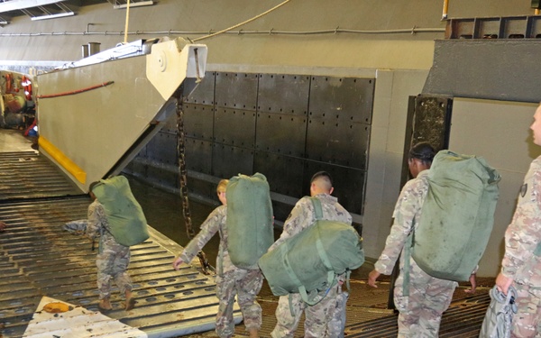 Military personnel in camouflage uniforms are boarding or loading onto what appears to be a naval vessel, likely a landing craft or ship's well deck. Soldiers are carrying large olive-drab duffel bags and rucksacks as they move in a line toward the vessel's ramp. The setting appears to be an amphibious military operation or troop embarkation exercise.