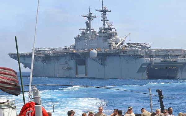 A large group of military personnel in camouflage uniforms are gathered on the deck of a naval vessel, with a large amphibious assault ship bearing the hull number '3' visible in the background at sea. An American flag is visible on the left side of the foreground vessel. The scene appears to depict a military operation or transit, likely involving a U.S. Navy amphibious ready group.