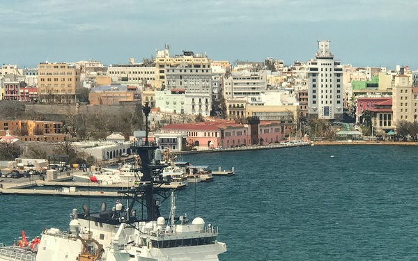 A U.S. Coast Guard cutter with hull number 754 is navigating through a harbor in front of a colorful coastal city, identifiable as San Juan, Puerto Rico. The white vessel displays the distinctive USCG diagonal red stripe and markings clearly on its hull. The urban waterfront backdrop features a mix of historic and modern architecture typical of Old San Juan.