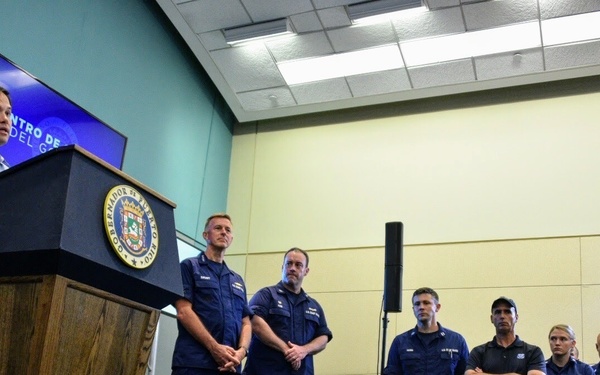 A man in civilian clothing speaks at a podium bearing an official government seal, addressing a group of uniformed personnel standing to his right. The setting appears to be an official press briefing or emergency response meeting, likely in Puerto Rico based on the visible flag and podium seal. Several individuals in navy blue uniforms, consistent with U.S. Coast Guard attire, are lined up attentively behind the speaker.