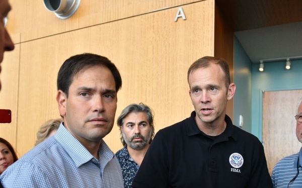 A group of individuals are gathered in what appears to be an indoor briefing or meeting setting. One person is wearing a black polo shirt with a visible 'FEMA' logo and Department of Homeland Security emblem, suggesting an emergency management context. Several people are engaged in conversation, and one person in the background is holding a red smartphone.