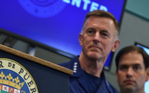 Two individuals stand at or near a podium bearing the official seal of the Governor of Puerto Rico ('Gobernador de Puerto Rico'). One person is wearing a U.S. Coast Guard uniform with visible rank insignia and a name tag partially reading 'ZUKUNFT' or similar. The setting appears to be an official press conference or briefing room with blue screens visible in the background.