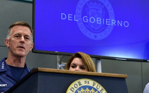 A press conference is being held at the Governor of Puerto Rico's podium ('Gobernador de Puerto Rico'). A military officer identified by his name tag as 'ZUKUNFT,' wearing a dark blue uniform with four-star insignia, is speaking at the podium. A woman stands behind the podium and a man in a blue shirt stands to the right, both observing the speaker.