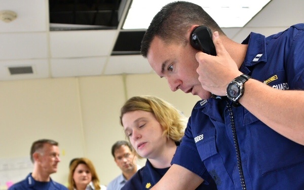 The image shows personnel in U.S. Coast Guard uniforms working in what appears to be an operations or command center. A male officer in the foreground is speaking on a corded telephone while looking at a computer monitor, alongside a female colleague who is interacting with the same screen. Several other uniformed and civilian personnel are visible in the background observing or discussing.