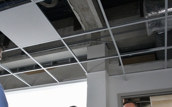 Two U.S. Coast Guard personnel in uniform are speaking with a civilian inside a damaged building. The ceiling shows significant structural damage with missing tiles, exposed ductwork, and visible cracks. The setting appears to be a post-disaster damage assessment or inspection.