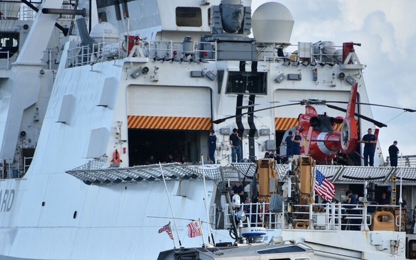 The image shows a U.S. Coast Guard small response boat moving at high speed in front of a large U.S. Coast Guard cutter. The large vessel, partially identified as 'JAMES' on the right side, has a red helicopter visible on its deck along with numerous personnel. The scene appears to be in a harbor or port area with overcast skies.