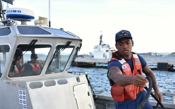 A U.S. Coast Guard crew member wearing an orange life vest and blue uniform is handling a mooring line on a small patrol boat numbered 35105. The scene appears to be at a Coast Guard dock or pier, with a larger Coast Guard cutter visible in the background on the water. Additional crew members are visible inside the boat's cabin.