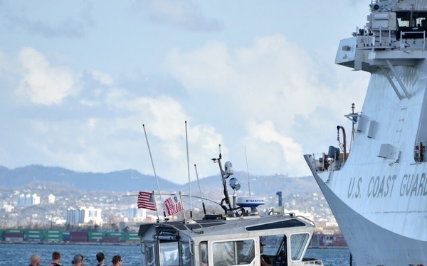 A U.S. Coast Guard response boat is underway at speed on open water, with several personnel seated in the rear cockpit area wearing life vests. The small orange and gray rigid inflatable boat (RIB) is operating in close proximity to a large U.S. Coast Guard cutter visible in the background. A cityscape and mountain range are visible in the far background, suggesting a harbor or port environment.
