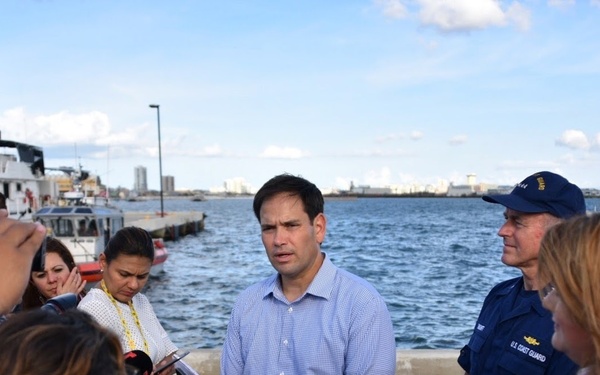 A man in a light blue checkered shirt stands at a waterfront dock speaking to a group of people, some holding phones and notepads, suggesting a press briefing or public statement. A person in a U.S. Coast Guard uniform stands nearby in the background. The setting appears to be a harbor or port facility with Coast Guard vessels visible.