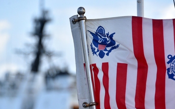 A United States Coast Guard ensign flag is displayed in sharp focus in the foreground, featuring the distinctive red and white vertical stripes, the Great Seal of the United States, and the USCG circular emblem dated 1790. In the blurred background, a large white Coast Guard cutter vessel is docked at what appears to be a harbor or port facility. The image uses a shallow depth-of-field technique to emphasize the flag while contextualizing it within a Coast Guard operational setting.