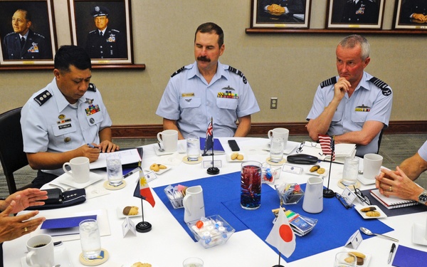 A formal military meeting is taking place around a round table with officers in light blue/grey uniforms from multiple nations. Small national flags including those of Japan, the Philippines, and what appears to be Australia or New Zealand are visible on the table, indicating a multinational military conference. The setting appears to be an official conference room with framed portraits of senior military officers on the walls.
