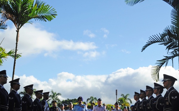 A formal military honor guard ceremony is taking place outdoors in a tropical setting, with two rows of uniformed personnel in dark dress uniforms lining a red carpet. Senior officers in light blue uniforms are walking down the center of the red carpet, suggesting a dignitary arrival or inspection. The setting features tall palm trees and a bright blue sky with scattered clouds.