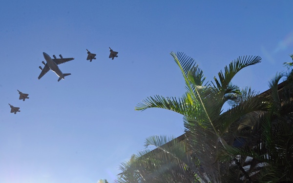 A military flyover formation is captured from a low ground-level perspective, showing one large transport or cargo aircraft flanked by four smaller fighter jets in a V-like formation against a clear blue sky. Palm trees and a building are visible on the right side, with at least one spectator looking upward. The image appears to be taken at a public event or ceremony in a tropical location.