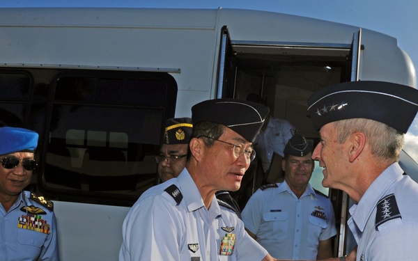 The image shows a formal military greeting between senior officers of what appear to be different air forces. Two officers in the foreground are shaking hands, while several other uniformed personnel stand nearby in front of a white transport van. The setting appears to be an outdoor tarmac or arrival area in a tropical location based on the vegetation and sky.