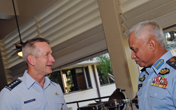 Two senior military officers in light blue uniforms are engaged in conversation at what appears to be a formal reception or dining event. The officer on the left wears a US Air Force uniform with three-star rank insignia, while the officer on the right displays a heavily decorated uniform with multiple medal ribbons and wing badges, suggesting a foreign air force. The setting appears to be an open-air venue with buffet tables visible in the background.