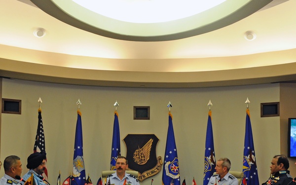 A formal military conference is taking place at a large oval conference table, identified by the screen display as the 'Pacific Air Chiefs Symposium 2017.' Multiple senior military officers from various nations are seated around the table, indicated by their diverse uniforms and rank insignia. The setting features a prominent Air Force emblem on the wall, multiple blue military flags, and national flag placards on the table.