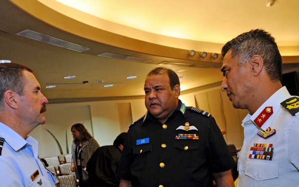 Three military officers in uniform are engaged in conversation at what appears to be a multinational military conference or meeting. The officer on the left wears a light blue uniform with an 'AUSTRALIA' shoulder patch, while the center officer wears a dark navy uniform with aviator wings and ribbons, and the right officer wears a white uniform with red rank insignia and a shoulder patch. The setting appears to be a hotel or conference facility with additional attendees visible in the background.