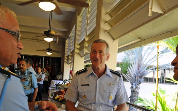 Three military officers in uniform are engaged in conversation around a white-clothed table at what appears to be a formal reception or dining event. The setting is an open-air covered venue with ceiling fans, suggesting a tropical or warm-climate location. Officers from at least two different nations appear to be present, indicated by differing uniform styles.