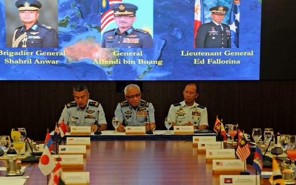 A multinational military conference is taking place around a large conference table, with three senior air force officers seated at the head table. A large display screen behind them shows portraits and names of three officials: Brigadier General Shahril Anwar, General Affendi bin Buang, and Lieutenant General Ed Fallorina. Multiple national flags are visible on the table, indicating representation from numerous countries across the Asia-Pacific region.