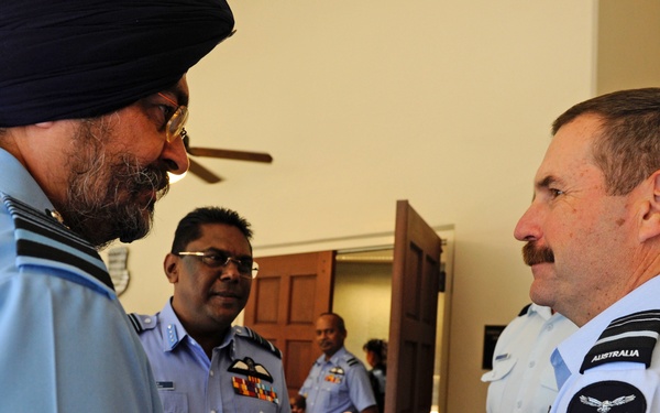 The image shows a group of military personnel in uniform engaged in conversation indoors. A man wearing a dark blue turban and light blue uniform is speaking with a man wearing an Australian Air Force uniform, identifiable by the 'Australia' and 'Air Force' shoulder patches. Additional uniformed personnel are visible in the background.