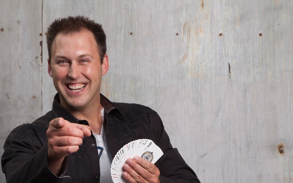 A smiling man stands in front of a weathered metal door, holding a fanned deck of playing cards in one hand while pointing toward the camera with the other. His expression and pose suggest he is a magician or card performer engaging with an audience. The image appears to be a professional or promotional photograph.