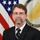 Douglas Lamont A formal official portrait of a middle-aged man wearing a dark suit with a black and white striped tie. He is posed in front of a United States flag and a partially visible government seal that reads 'Senior Executive Service.' The image appears to be a standard U.S. government or military civilian official headshot.